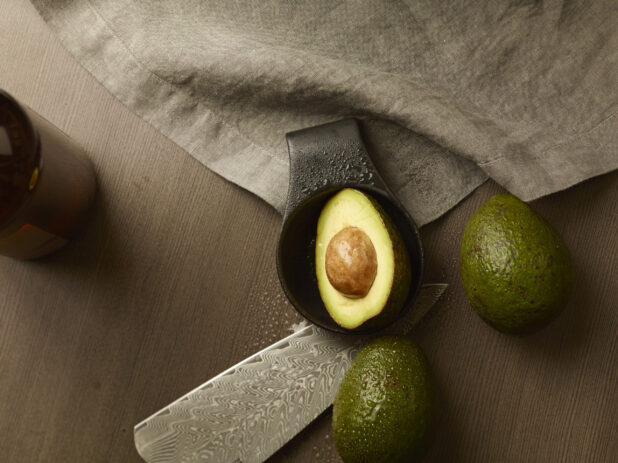 Overhead of an avocado cut in half in a dish with two other avocados and a knife around it on a wood table