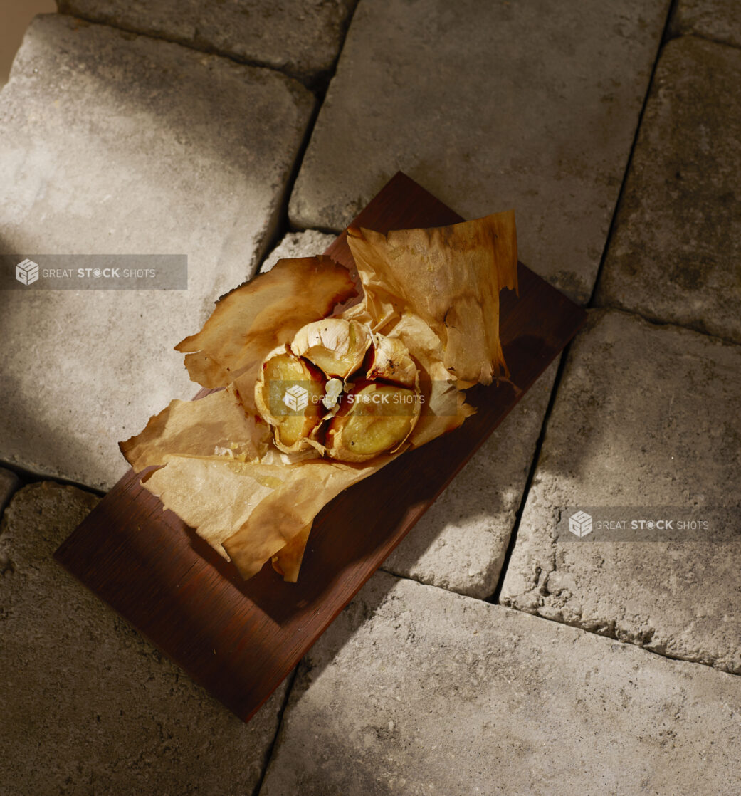 Overhead of a bulb of roasted garlic on parchment paper on a wooden board on stone