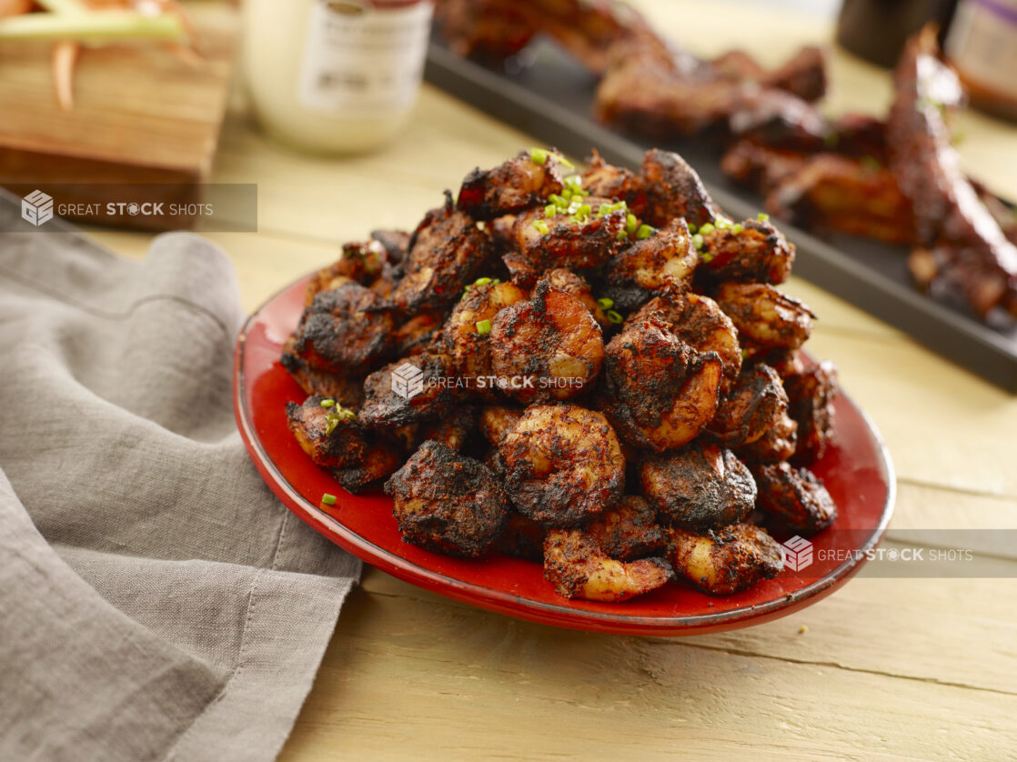 Red bowl of blackened shrimp topped with chives on a wood table with a platter of ribs in the background