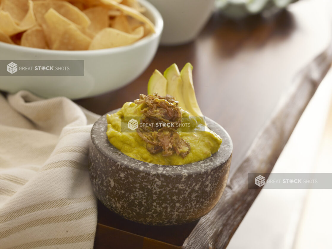 Small granite bowl of guacamole with sliced avocado and fried jalapeño with a bowl of tortillas in the background on a live edge wood table