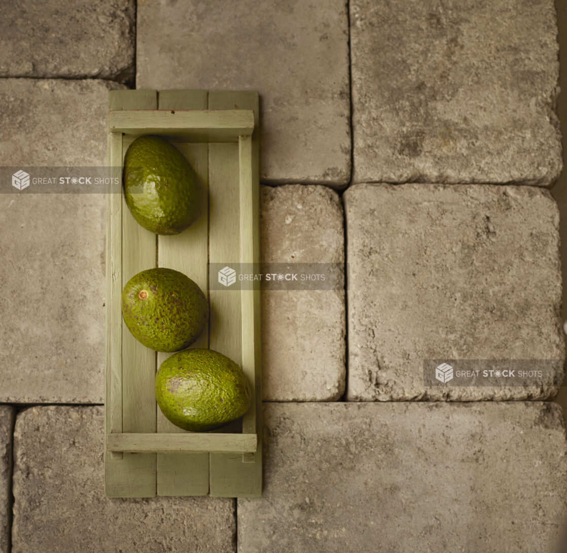 Overhead of three green avocados in a wood basket on stone