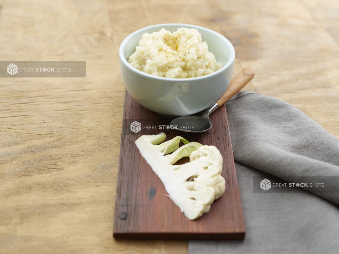 Bowl of cauliflower mash with a slice of fresh cauliflower on a wood board with a napkin and spoon on a wood table