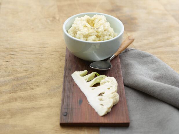 Bowl of cauliflower mash with a slice of fresh cauliflower on a wood board with a napkin and spoon on a wood table