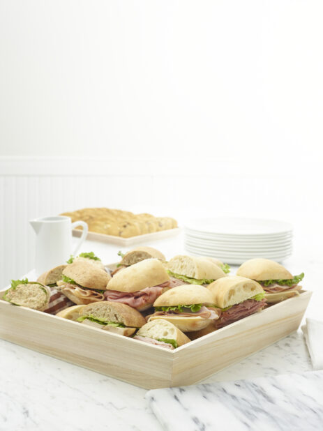Wooden tray of assorted deli sandwiches on a white marble table with a tray of cookies in the background
