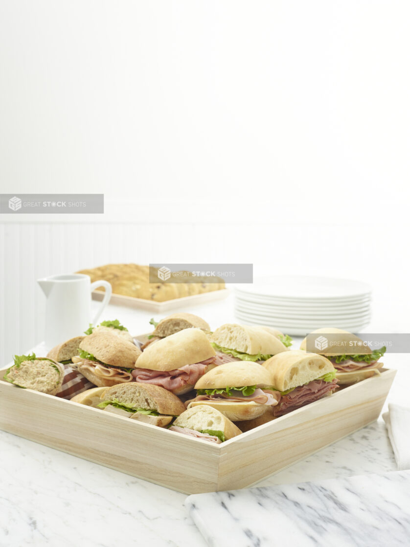 Wooden tray of assorted deli sandwiches on a white marble table with a tray of cookies in the background