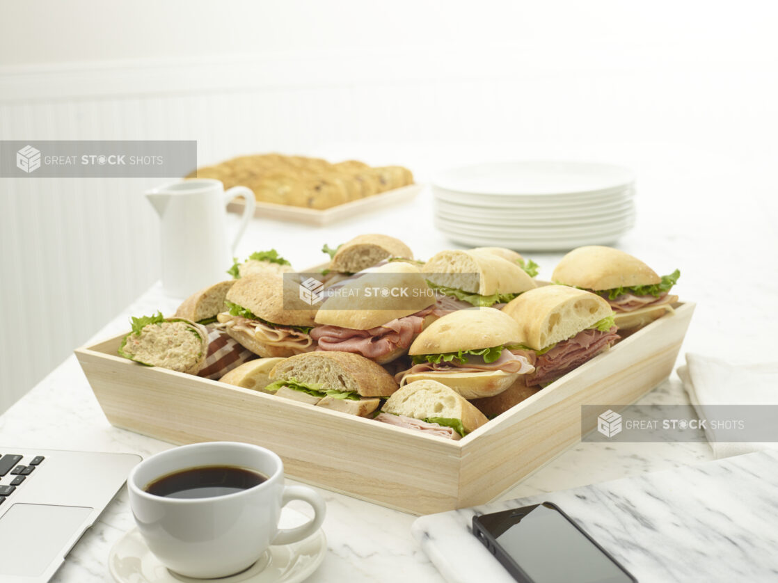 Wooden tray of assorted deli sandwiches on a white marble table with a laptop and cell phone in the background