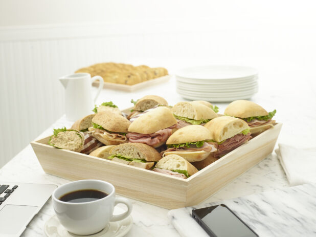 Wooden tray of assorted deli sandwiches on a white marble table with a laptop and cell phone in the background