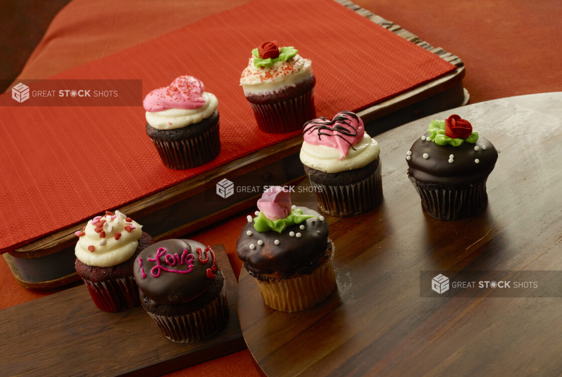 Valentine’s Day cupcakes decorated with roses, handwriting and hearts in chocolate, vanilla and red velvet with frosting on a wood table