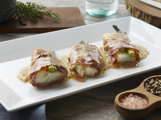 Three prosciutto wrapped white fish portions with asparagus on top of sauce on a white plate