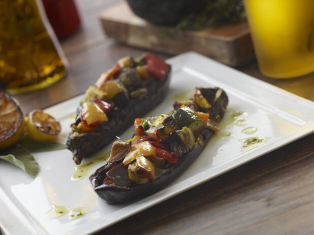 Two baked eggplant halves stuffed with peppers, eggplant and zucchini on a white plate drizzled in vinaigrette with grilled fresh lemon halves on a wood table