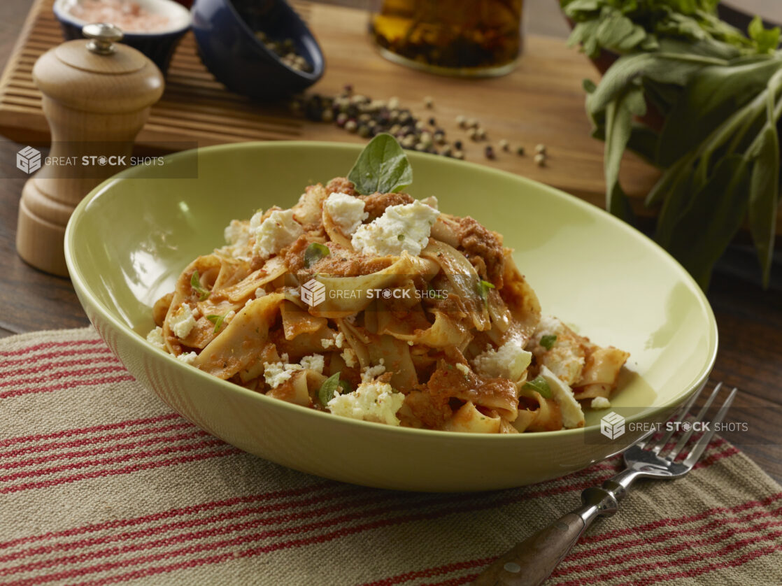 Large bowl of pappardelle pasta with a bolognese sauce, ricotta cheese and fresh basil in a green bowl with seasonins in the background