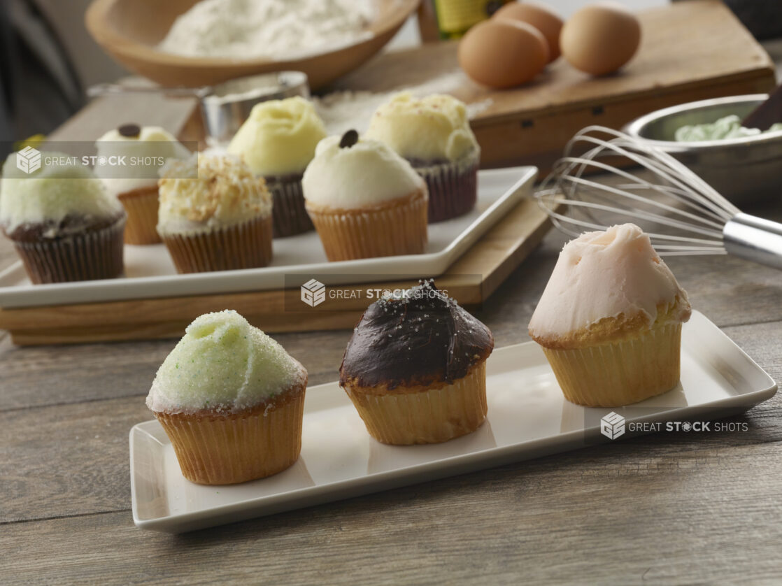 Assortment of different types of iced cupcakes on white rectangular plates with ingredients in the background on a wood table