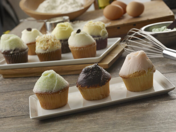Assortment of different types of iced cupcakes on white rectangular plates with ingredients in the background on a wood table