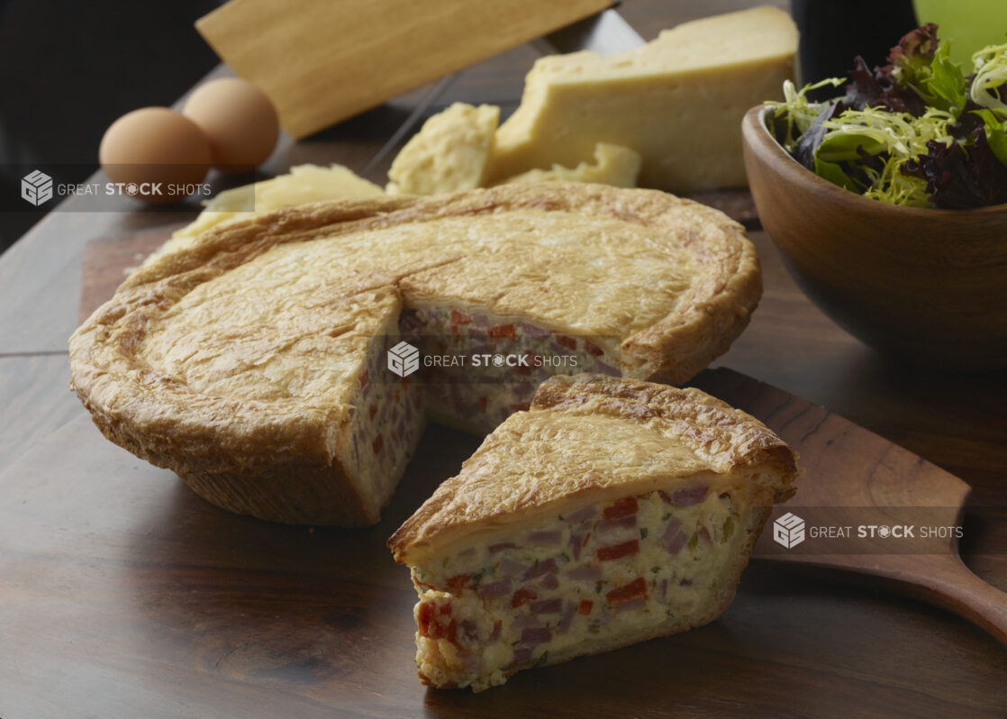 Slice of pizza rustica with ham, red pepper, cheese and green onion in a golden pie crust with the whole pie in the background and a side salad, whole eggs and a block of fresh cheese on a wood table