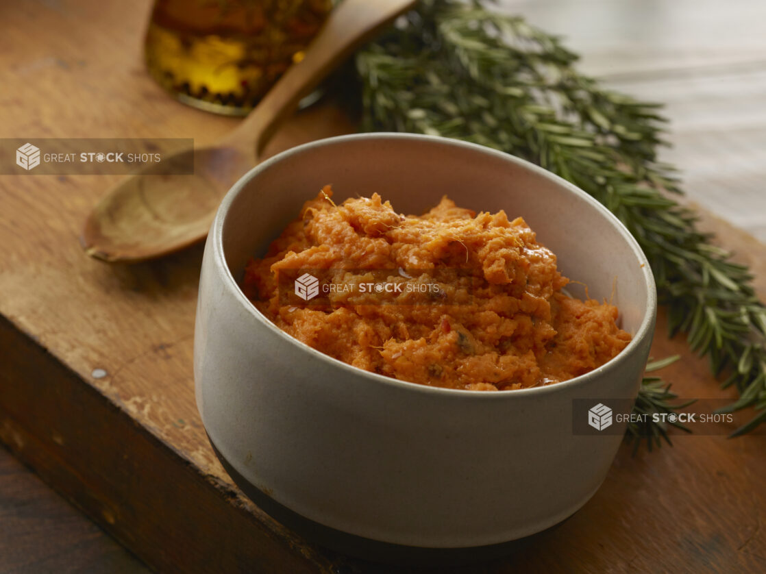 Mashed sweet potato in a straight-sided grey bowl with a wooden spoon and fresh rosemary in the background on a wood block