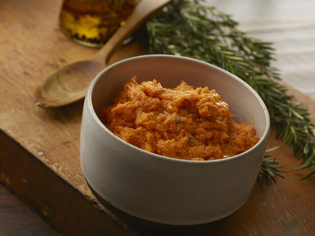 Mashed sweet potato in a straight-sided grey bowl with a wooden spoon and fresh rosemary in the background on a wood block