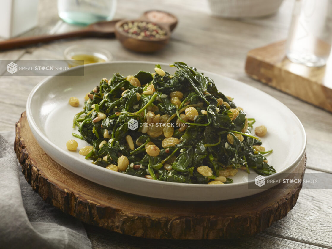 Warm wilted spinach salad with golden raisins and toasted pine nuts tossed in a garlic and olive oil dressing in a flat bottom bowl on a live edge wood medallion