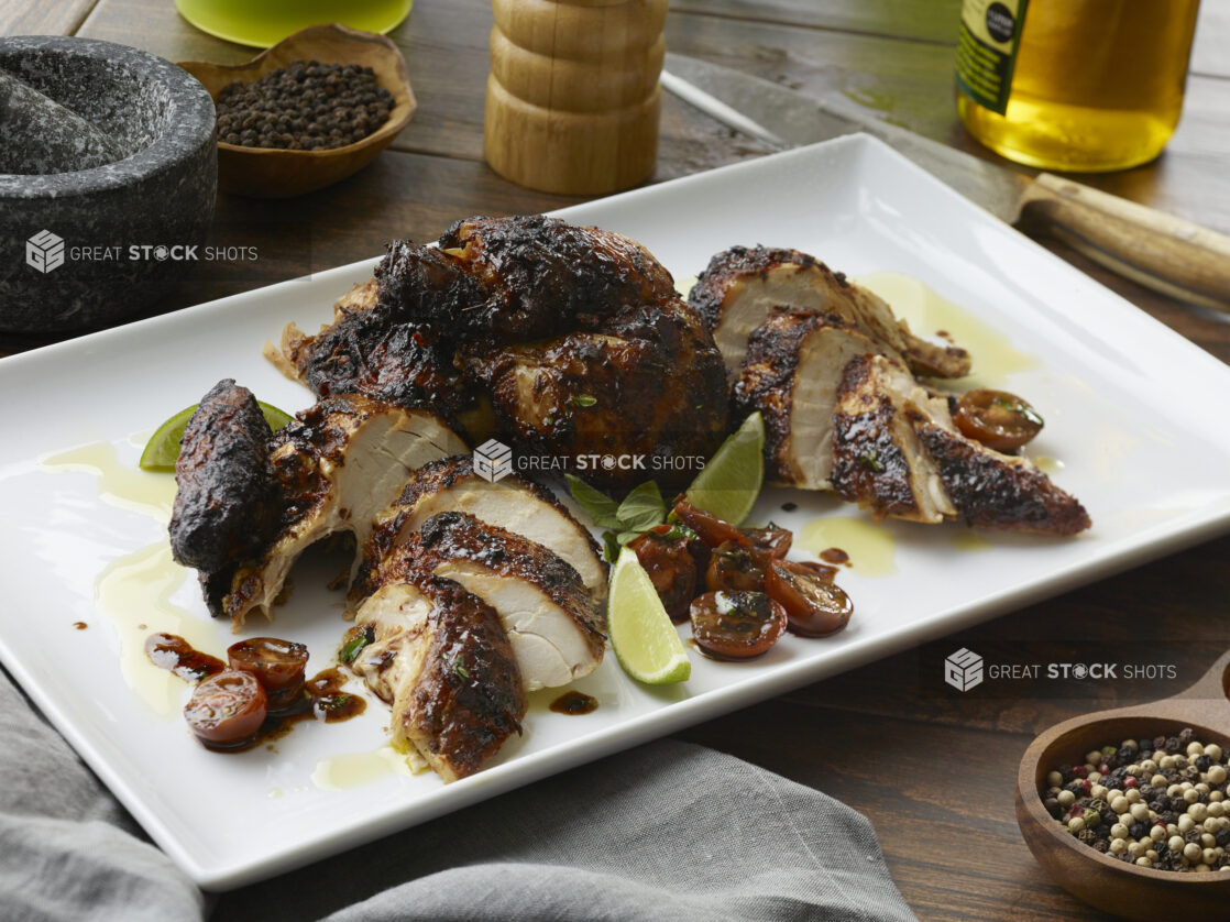 Sliced whole blackened balsamic chicken with halved cherry tomatoes and lime wedges on a white platter on a wood table