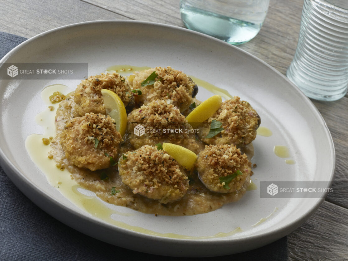 Crumb topped baked clams on top of sauce with lemon wedges and parsley, drizzled with olive oil on a white plate