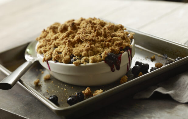 White baking dish of blueberry crumble on a baking sheet with a spoon