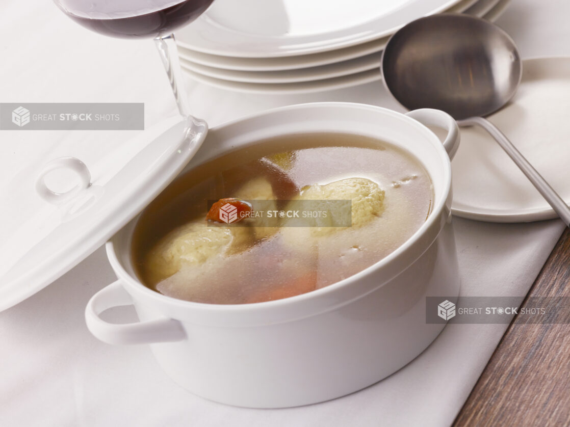 Small ceramic bowl of matzo ball soup with lid on a white table cloth with ladle and red wine in the background