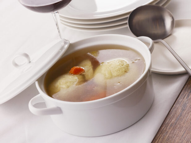 Small ceramic bowl of matzo ball soup with lid on a white table cloth with ladle and red wine in the background