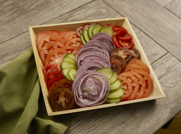 Wood catering tay with a variety of sliced tomatoes, red onion and cucumbers on a wood table with a napkin
