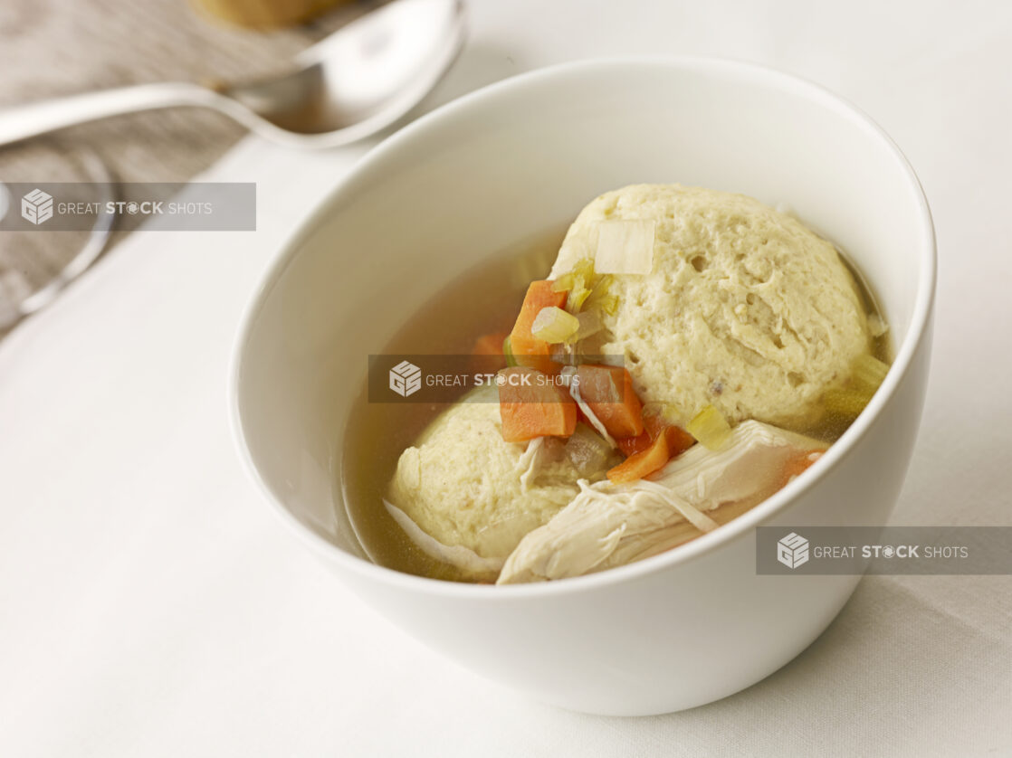 Small white bowl of matzo ball soup on a white table cloth with a spoon in the background