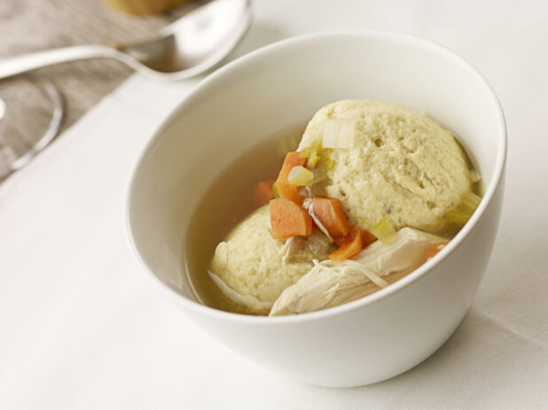 Small white bowl of matzo ball soup on a white table cloth with a spoon in the background