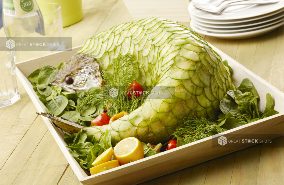 Catering presentation of cold poached salmon covered in cucumber scales on a wood catering tray with spinach, fresh lemon and fresh dill