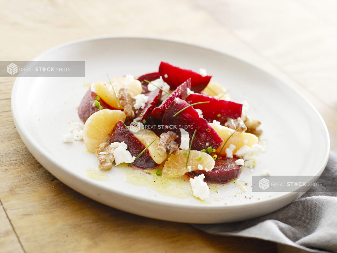 Large white plate with sliced beets, oranges, goat cheese and walnuts garnished with chives on a wood table