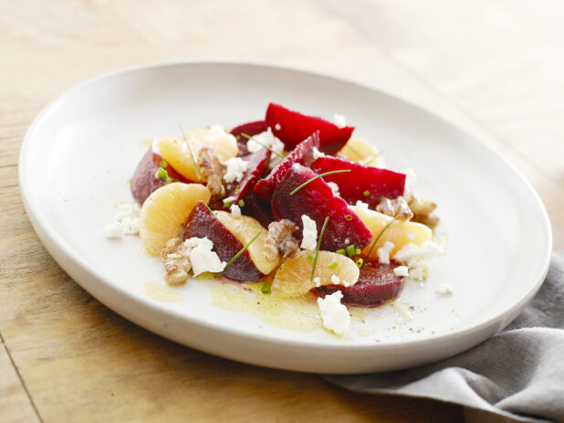 Large white plate with sliced beets, oranges, goat cheese and walnuts garnished with chives on a wood table