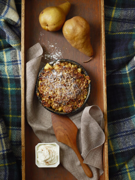Overhead baked pear crisp dessert topped with icing sugar with two fresh pears and a side dish of vanilla ice cream on a wood tray with a tartan background
