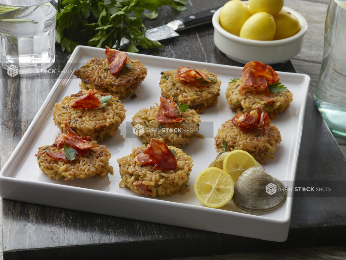 Fried oyster rice cakes topped with a pancetta chip and fresh parsley on a white platter with fresh lemon