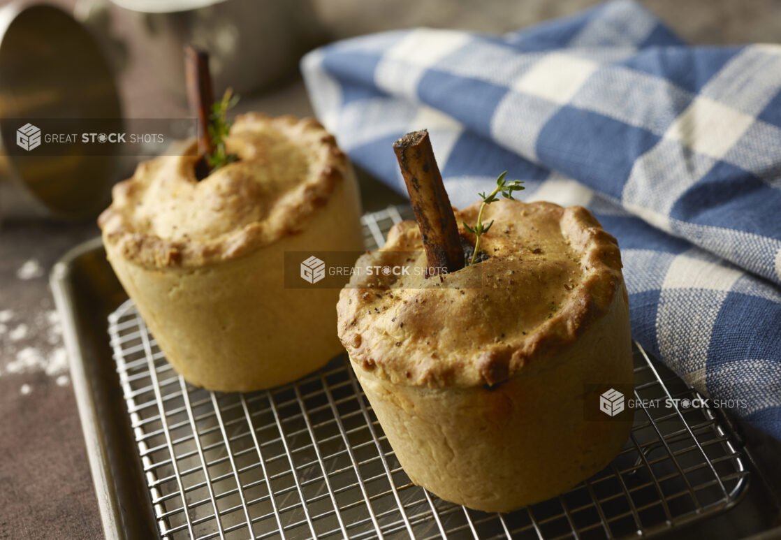 Two standing beef pies with bone in the middle and fresh herbs on a cooling rack in a kitchen with a checkered napkin