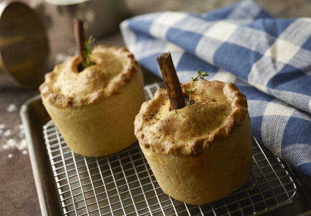 Two standing beef pies with bone in the middle and fresh herbs on a cooling rack in a kitchen with a checkered napkin