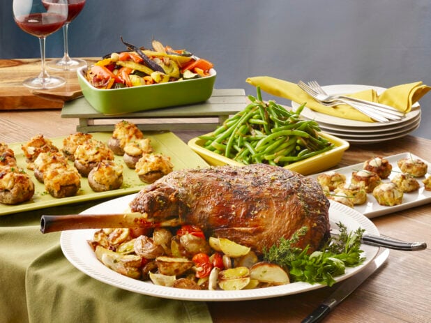 Large feast of bone in prime rib roast with roasted potatoes and cherry tomatoes, sides of green beans and roasted vegetables and appetizers on a wood table