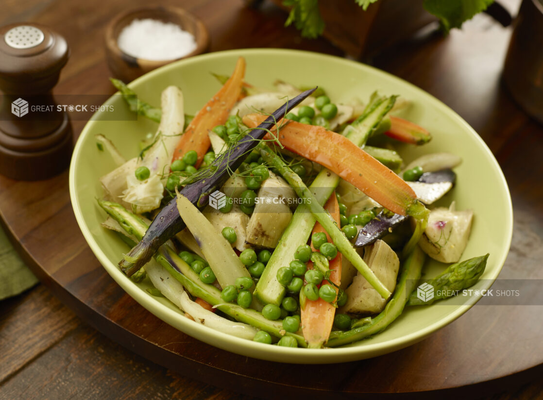 Green bowl of steamed rainbow carrots, asparagus, artichoke and peas on a wooden board