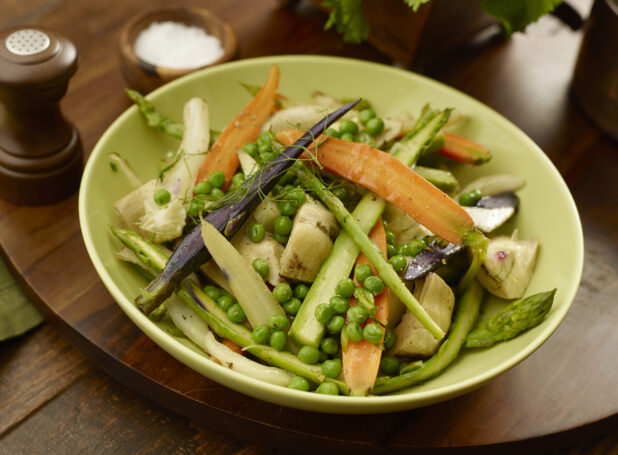 Green bowl of steamed rainbow carrots, asparagus, artichoke and peas on a wooden board