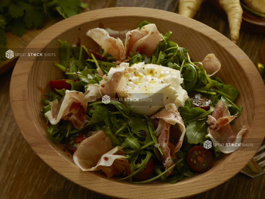 Burrata chees on top of an arugula salad with prosciutto and cherry tomatoes, drizzled in olive oil and a balsamic vinaigrette in a wooden bowl