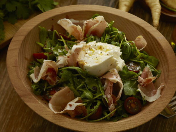 Burrata chees on top of an arugula salad with prosciutto and cherry tomatoes, drizzled in olive oil and a balsamic vinaigrette in a wooden bowl