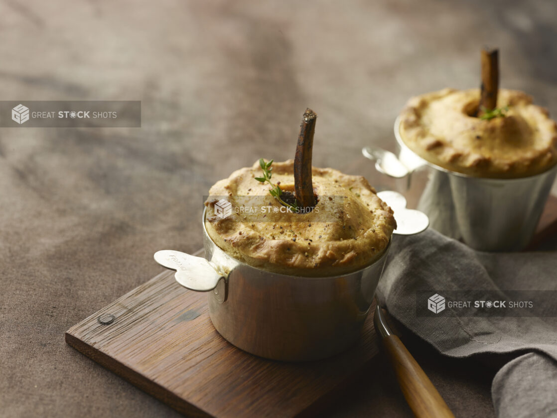 Two beef pot pies with bone in the middle and fresh herbs in a metal pot on a wooden board