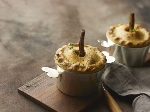 Two beef pot pies with bone in the middle and fresh herbs in a metal pot on a wooden board