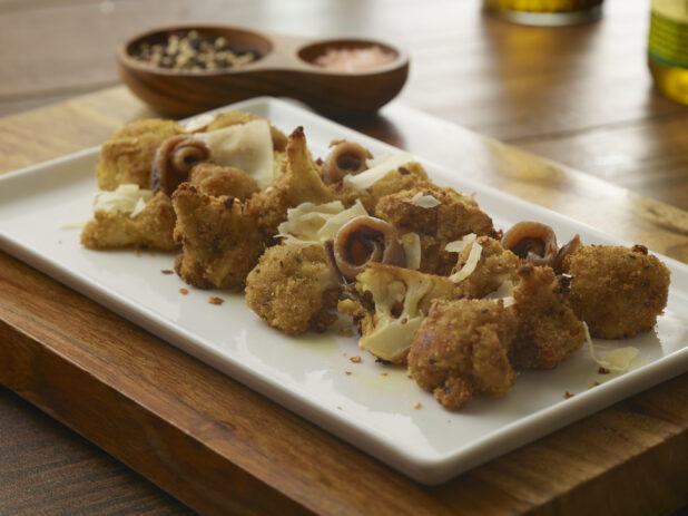 Appetizer platter of fried cauliflower bites and rolled anchovies garnished with shaved parmesan on a white rectangular platter on a wooden board on a wood tabletop