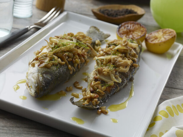 Two whole roasted branzino fish topped with fennel, brown butter and toasted pine nuts on a white dish with charred lemon halves on a wood table