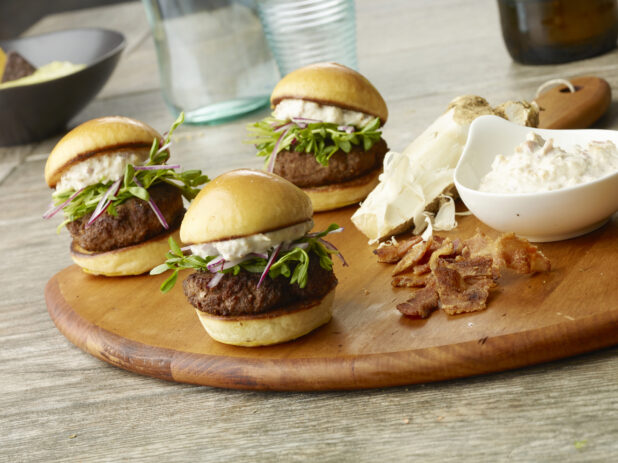 Three hamburger sliders with microgreens, red onion and a creamy bacon, cheese sauce on a toasted bun on a wooden board