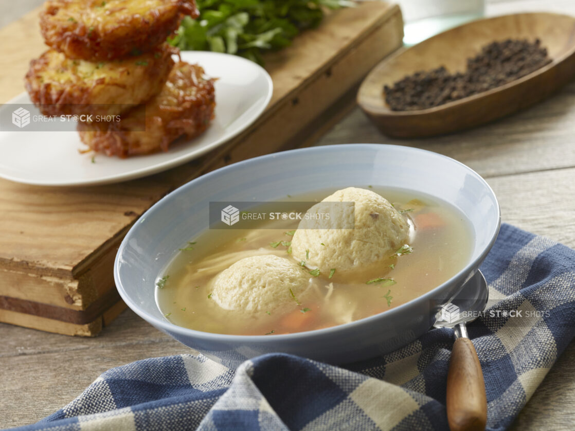 Light blue bowl of matzo ball soup with potato latkes in the background on a wooden board with fresh parsley