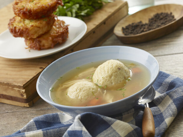 Light blue bowl of matzo ball soup with potato latkes in the background on a wooden board with fresh parsley