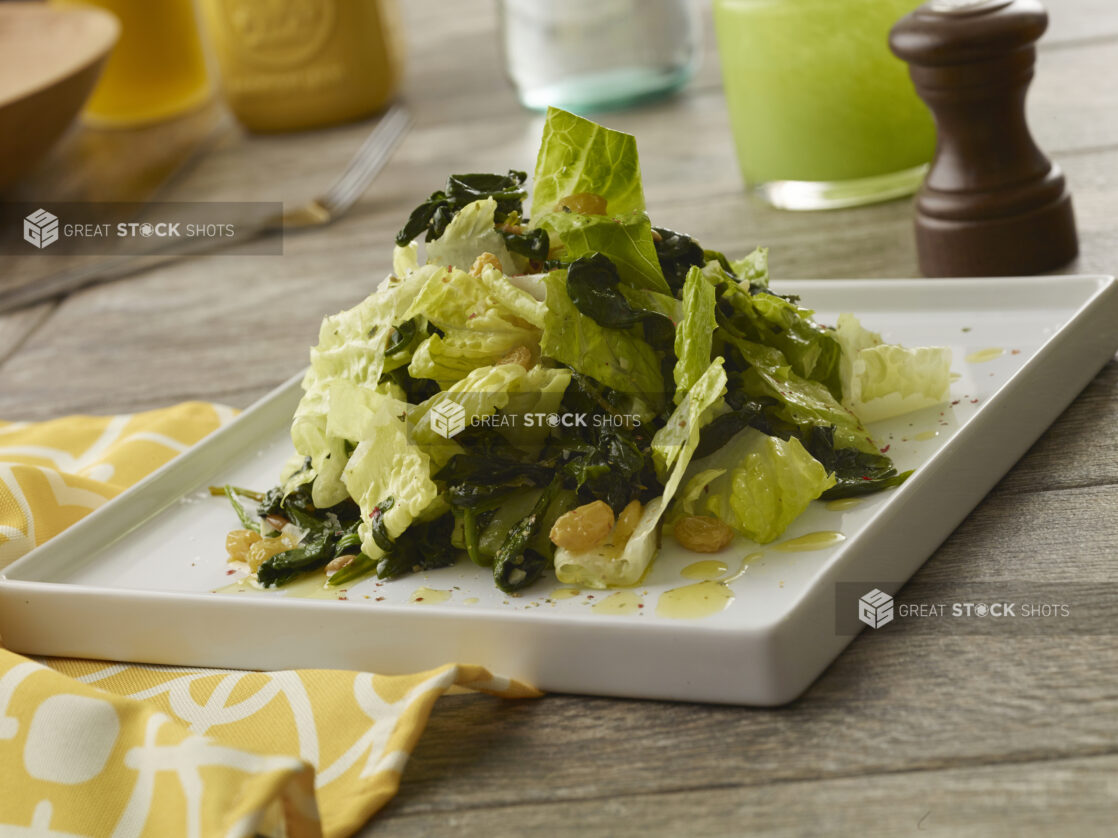 Small white square plate of leaf salad with golden raisins tossed in a vinaigrette dressing on a wood table