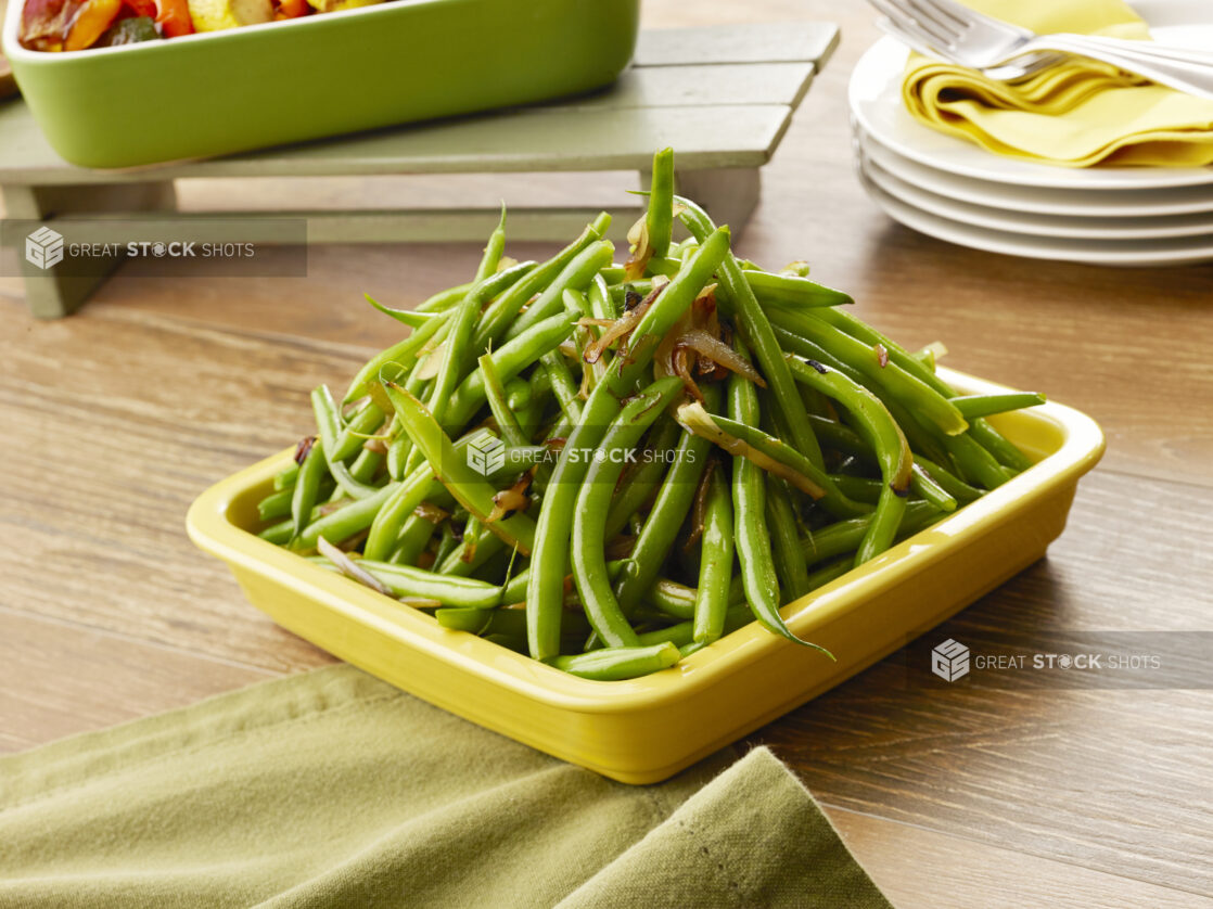 Yellow bowl of cooked green beans with caramelized onions on a wood table